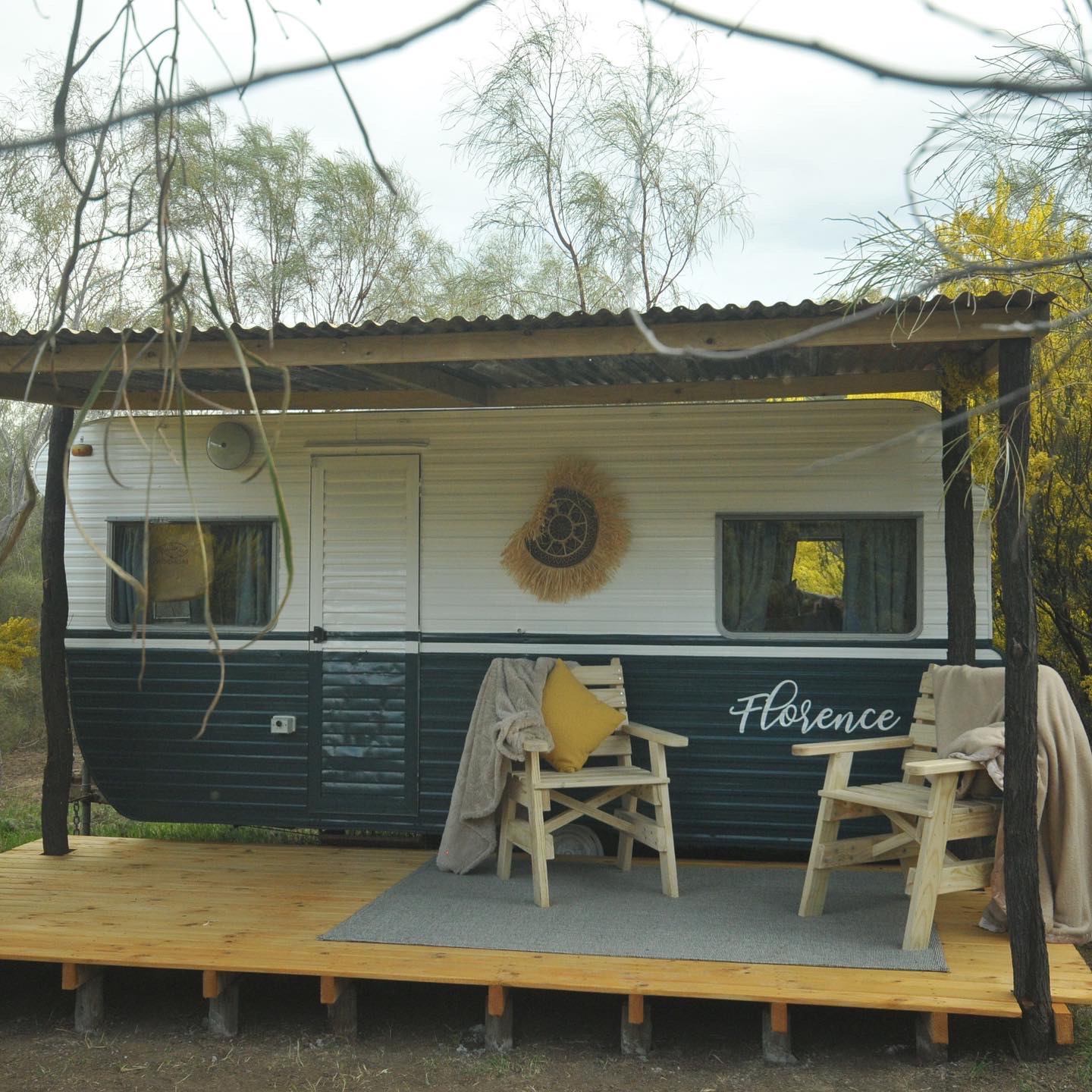 Florence, the vintage caravan, painted dark green and cream under a corrugated iron shelter with two deck chairs on the timber deck